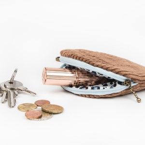 May include: A brown corduroy coin purse with a light blue lining and a leopard print interior. The purse is open and contains a rose gold lipstick and a set of keys. There are also several euro coins scattered on the white surface.