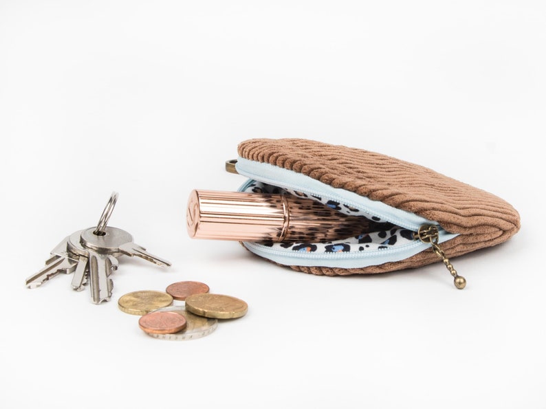 May include: A brown corduroy coin purse with a light blue lining and a leopard print interior. The purse is open and contains a rose gold lipstick and a set of keys. There are also several euro coins scattered on the white surface.