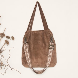 May include: A brown corduroy tote bag with a white patterned strap. The bag has a top handle and a long adjustable strap.