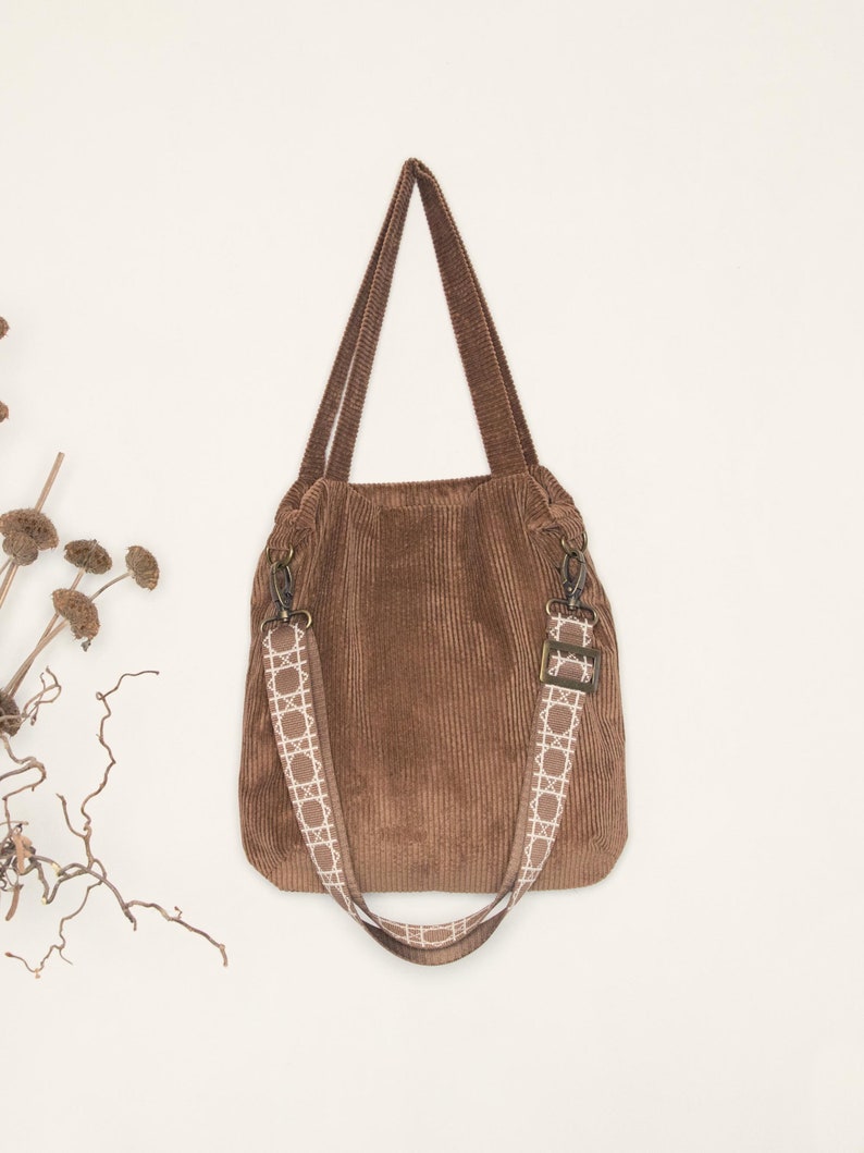 May include: A brown corduroy tote bag with a white patterned strap. The bag has a top handle and a long adjustable strap.
