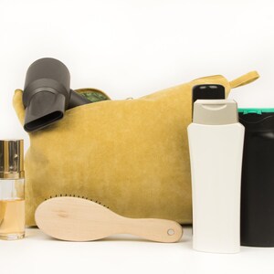 May include: A mustard yellow corduroy cosmetic bag with a black hairdryer, a wooden hairbrush, two bottles, and a perfume bottle. The bottles are white and black, and the perfume bottle is clear with a gold cap.