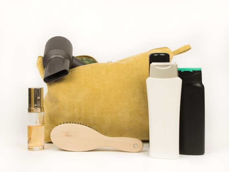 May include: A mustard yellow corduroy cosmetic bag with a black hairdryer, a wooden hairbrush, two bottles, and a perfume bottle. The bottles are white and black, and the perfume bottle is clear with a gold cap.