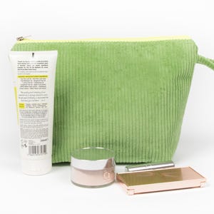 May include: A green corduroy cosmetic bag with a yellow zip, a tube of lotion, a jar of pink cream, a lipstick, and a rose gold compact. The lotion tube has text on it. The items are arranged on a white surface.