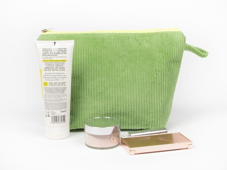 May include: A green corduroy cosmetic bag with a yellow zip, a tube of lotion, a jar of pink cream, a lipstick, and a rose gold compact. The lotion tube has text on it. The items are arranged on a white surface.