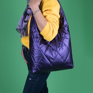 Peut inclure: Un sac fourre-tout matelassé violet foncé avec un motif en losanges. Le sac a une finition brillante et métallique et une apparence douce et rembourrée. Le sac est porté par une personne vêtue d'un sweat-shirt jaune et d'un jean bleu.