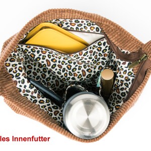 May include: A brown corduroy tote bag with a zipper closure. The bag is open and the interior is lined with a white fabric with a leopard print. The bag contains a yellow folder, a black phone, a silver water bottle, and a brown makeup stick.