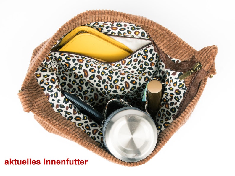May include: A brown corduroy tote bag with a zipper closure. The bag is open and the interior is lined with a white fabric with a leopard print. The bag contains a yellow folder, a black phone, a silver water bottle, and a brown makeup stick.