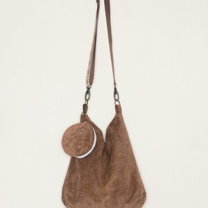 May include: A brown corduroy tote bag with a matching brown corduroy pouch attached to the front. The bag has a long adjustable strap.
