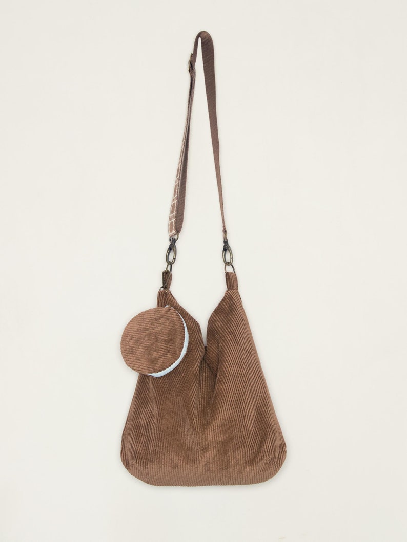 May include: A brown corduroy tote bag with a matching brown corduroy pouch attached to the front. The bag has a long adjustable strap.