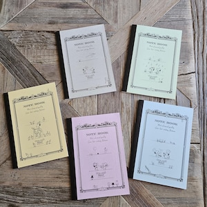 Op de afbeelding: Vijf rechthoekige notitieboeken met zwarte ruggen en pastelkleurige covers in geel, grijs, lichtgroen, roze en lichtblauw. Elke cover heeft een decoratieve zwarte rand, de tekst NOTE BOOK en kleine zwarte lijntekeningen van Peanuts-personages.