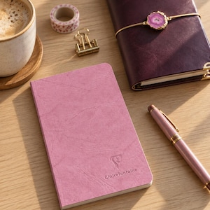 Puede incluir: Un cuaderno rosa Clairefontaine con cubierta texturizada, un diario morado oscuro con cierre de piedra de ágata rosa, un bolígrafo rosa, un pequeño rollo de cinta adhesiva decorativa, un clip de metal dorado y una taza de cerámica con café.