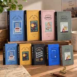 Puede incluir: Una colección de nueve cuadernos de viaje rectangulares en varios colores, incluyendo azul, amarillo, rosa, verde y negro. Cada cuaderno presenta ilustraciones gráficas únicas, texto y lomos negros. Los cuadernos están dispuestos en dos filas.