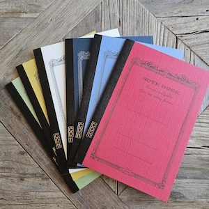 Puede incluir: Un conjunto de seis cuadernos dispuestos en abanico, con cubiertas texturizadas en rojo, azul claro, azul oscuro, blanco, amarillo y verde claro. Cada cuaderno tiene un lomo negro, un borde decorativo negro y el texto NOTE BOOK CD 11.