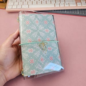 Puede incluir: Un cuaderno con una cubierta decorativa con un estampado floral en azul claro y rosa. El cuaderno se guarda en una funda de plástico transparente y se asegura con un lazo dorado y una banda elástica verde menta.