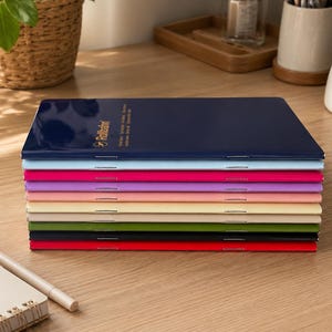 May include: A stack of nine slim, rectangular notebooks with stapled spines, arranged in a vertical pile. The top cover is dark blue with the text Rollbahn printed in gold. The remaining notebooks feature solid colours including light blue, pink, purple, light pink, cream, beige, green, and red.