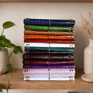 Puede incluir: Una pila vertical de doce diarios rectangulares con un acabado texturizado de imitación de piel de cocodrilo en varios colores, incluyendo azul marino, verde, tostado, naranja, rojo, blanco, verde azulado, marrón oscuro, burdeos, morado y rosa claro.