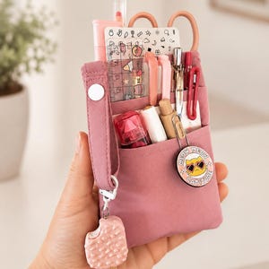 Puede incluir: Un estuche de tela color rosa empolvado con varios bolsillos que contiene bolígrafos, una regla transparente y tijeras. Un dije de helado rosa cuelga de una correa lateral, junto a un pin redondo con un gato con gafas de sol en forma de corazón.