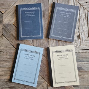 Puede incluir: Cuatro cuadernos rectangulares con lomos negros y cubiertas texturizadas en azul oscuro, gris, azul claro y crema. Cada cubierta presenta un marco negro decorativo, el texto "NOTE BOOK", "Most advanced quality" y "Good best writing features".