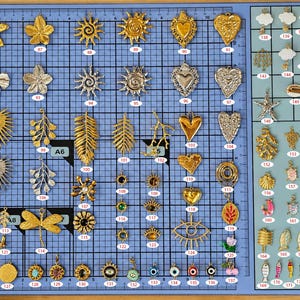 Könnte beinhalten: Eine Sammlung verschiedener Metallanhänger, die auf einer blauen Rastermatte angeordnet sind. Die Anhänger haben verschiedene Formen wie Blumen, Sonnen, Herzen, Blätter, Sterne, Tiere und Augen, jeweils mit einem nummerierten Etikett versehen.
