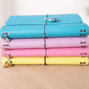 May include: A stack of four colorful journals in shades of blue, lavender, pink, and yellow. Each journal is bound with a leather cord and has decorative charms. The journals are stacked on a light wooden surface.
