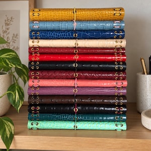 Puede incluir: Una pila vertical de catorce diarios rectangulares con cubiertas texturizadas de piel de reptil sintética en varios colores, incluyendo amarillo, azul, rojo, negro, morado y turquesa. Cada diario tiene ojales de metal dorado y un cierre elástico.