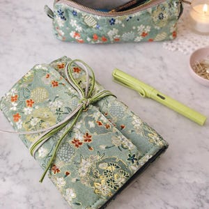 Op de afbeelding: Een groene etui met bloemenpatroon en een bijpassende oprolbare pennenzak, vastgebonden met een groen en wit lint. Een limoengroene pen en een etui met rits met pennen erin zijn ook zichtbaar.