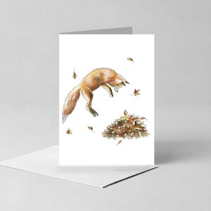 May include: A greeting card featuring a watercolor illustration of an orange fox mid-leap, pouncing toward a pile of brown and yellow autumn leaves. Several individual fallen leaves are scattered around the fox against a plain white background.