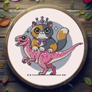 May include: Cross-stitch design featuring a cat wearing a crown and a bow tie, riding a pink dinosaur. The cat has orange, gray, and white fur. The dinosaur is pink. The design is set in a wooden embroidery hoop.