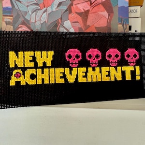 Peut inclure: Une œuvre d'art en point de croix noire avec les mots "NEW ACHIEVEMENT!" en jaune et quatre crânes roses. L'œuvre rectangulaire semble faite à la main, avec une texture en forme de grille.
