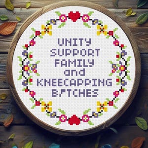 May include: A circular embroidery hoop containing white fabric with a cross-stitch design. The text reads UNITY SUPPORT FAMILY and KNEECAPPING B/TCHES in purple. A floral wreath with red hearts, yellow, pink, and purple flowers surrounds the text.