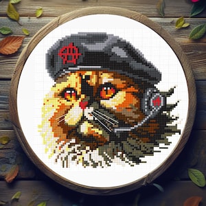 May include: Cross-stitch image of a cat wearing a black beret with a red emblem and a headset. The cat has orange and brown fur, with detailed stitching creating a pixelated effect. The artwork is set in a wooden embroidery hoop.