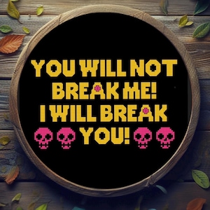 May include: A black cross-stitch design in a wooden hoop features the text "YOU WILL NOT BREAK ME! I WILL BREAK YOU!" in yellow pixelated letters. Pink pixelated skulls are at the bottom. The background is a wooden surface with leaves and a rose.