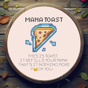 May include: A cross-stitch design on a round wooden frame. The design features the text "MANA TOAST" above a pixelated toast graphic. Below the graphic, text reads "THIS IS TOAST. IT REFILLS YOUR MANA. THAT'S IT NOTHING MORE. FOCK YOU."