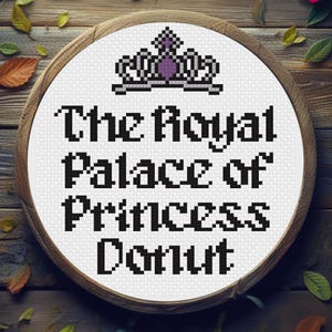 May include: A cross-stitch design in a wooden hoop features the text "The Royal Palace of Princess Donut" in black pixelated letters. A pixelated crown is above the text. The background is a wooden surface with scattered leaves and a rose.