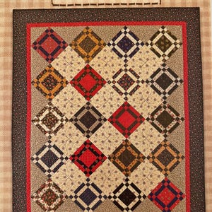 May include: A quilt featuring a grid of diamond-shaped fabric patches in red, yellow, blue, and floral patterns, bordered by a red and black frame. The text Heartspun Quilts and Peacefield is printed on the beige checkered background.
