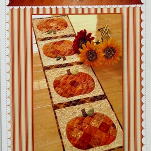 May include: A rectangular patchwork table runner featuring four square blocks, each displaying a quilted orange pumpkin with green stems on a cream background. The text Shabby Fabrics and Patchwork Pumpkin Table Runner is printed on the packaging.
