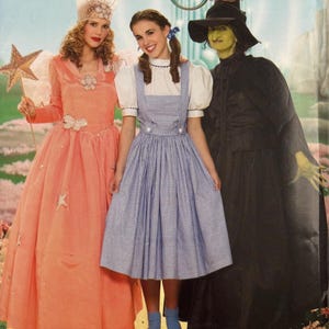 May include: A Simplicity sewing pattern envelope for The Wizard of Oz costumes, featuring three outfits: a peach-colored gown with a star wand, a blue and white gingham pinafore dress with a white blouse, and a black witch costume with a pointed hat.