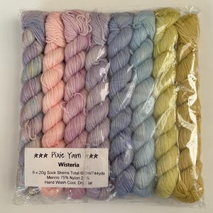May include: Eight skeins of yarn in a clear plastic bag, arranged in a gradient from light purple to pink, lavender, blue, and yellow-green. A white label reads: Pixie Yarn Wisteria, 8 x 20g Sock Skeins Total 680m/744yds, Merino 75% Nylon 25%, Hand Wash Cool, Dry Flat.