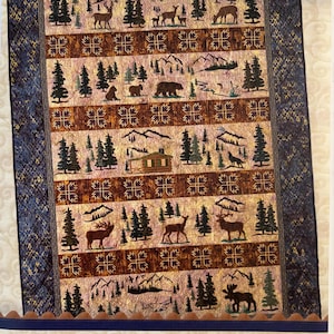 May include: A quilt pattern booklet titled Lunch Box Quilts Wilderness Wildlife Classic Series. The cover features a rectangular quilt design with rows of brown and tan fabric blocks depicting deer, bears, mountains, pine trees, and a cabin.