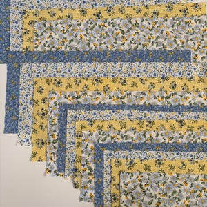 May include: A collection of fabric squares in a variety of floral patterns. The fabrics feature blue, yellow, and white daisy and floral designs. The squares are arranged in a layered, staggered pattern.