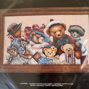 May include: Cross-stitch kit featuring a group of teddy bears wearing hats and outfits, framed in a wooden border. The bears are in various colors, including brown, blue, and pink. The kit includes 14-count cotton Aida fabric, 6-strand cotton floss, beads, needles, graph, and instructions.