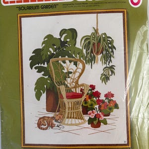 May include: A Sunset Stitchery Solarium Garden embroidery kit package featuring a printed design of a rattan chair, a hanging plant, potted flowers, and a cat on a tiled floor. The packaging indicates it is for a 16 inch by 20 inch frame.