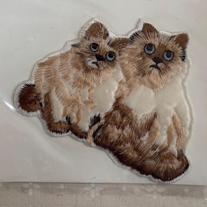 May include: An embroidered patch featuring two fluffy cats with light brown and cream fur, dark brown ears and tails, and bright blue eyes. The cats are positioned side by side, rendered in detailed thread work with a white border outline.