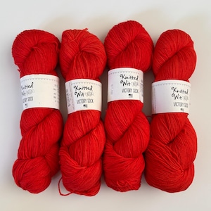 May include: Four skeins of bright red yarn arranged side by side. Each skein features a white paper label with black text that reads Knitted Wit and VICTORY SOCK, accompanied by a small candy graphic and a tiny American flag icon.