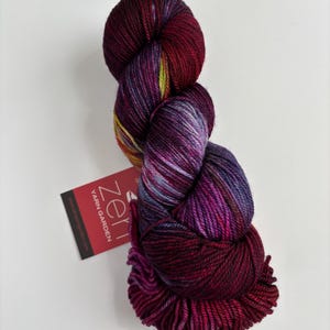 May include: A skein of hand-dyed yarn featuring a variegated color palette of deep burgundy, magenta, violet, and subtle hints of lime green. A rectangular red tag attached to the yarn displays the white text Zen YARN GARDEN.