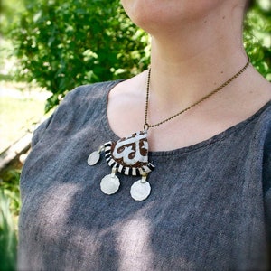 May include: A silver necklace with a brown and white fabric pendant featuring a geometric design. The pendant has four silver circles hanging from it.
