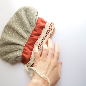 May include: A gray and orange fabric hat with white lace trim. The hat is made of a soft, textured fabric and has a gathered design.