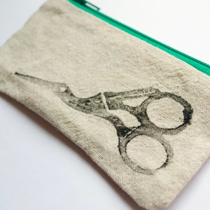 May include: A small, beige zippered pouch with a green zipper. The pouch has a black and white printed image of a pair of scissors.
