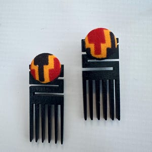 May include: A pair of earrings featuring black wooden comb-shaped pendants topped with circular fabric buttons. The fabric displays a geometric pattern in red, yellow, and black. The earrings have a long, rectangular design with four vertical prongs.