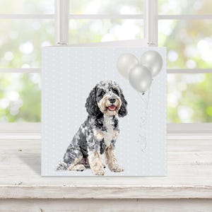 May include: A square greeting card featuring a watercolour illustration of a dog with black and grey fur, sitting with a light blue background with white polka dots. Three silver balloons are tied to the card.